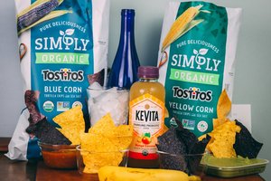 Two bags of Simply Organic Tostitos chips (blue and yellow corn) are displayed with bowls of chips, salsa, and guacamole. A Kevita Mango Coconut drink, a blue bottle, a glass of ice, and a banana accompany the setup on a wooden surface.